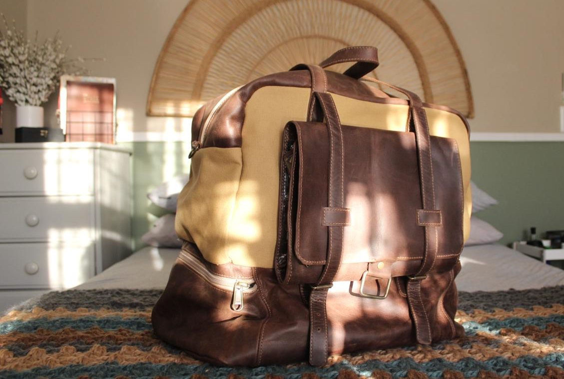 A beige and brown leather travel bag with a shoe pocket, placed on a bed