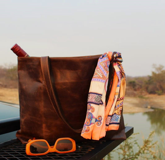A brown leather tote bag with a multicolored scarf and orange sunglasses placed on a surface, possibly wood, with a natural landscape in the background.