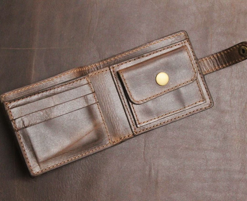 A brown, handmade leather wallet with a secure coin pocket, multiple card slots, and a visible brand emblem, placed on a dark surface