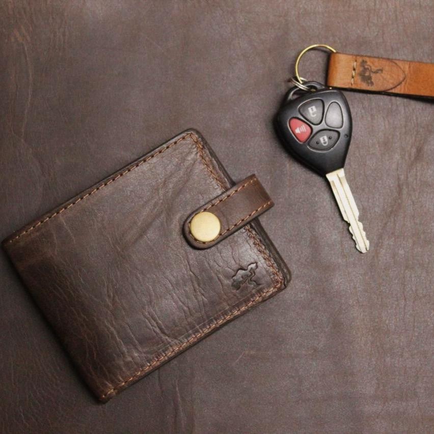 A brown, handmade leather wallet with a secure coin pocket, multiple card slots, and a visible brand emblem, placed on a dark surface next to a set of car keys