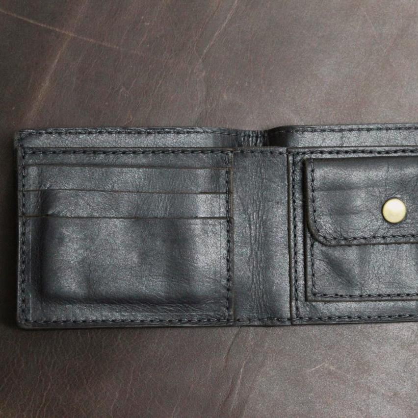 A black, handmade leather wallet with a secure coin pocket, multiple card slots, and a visible brand emblem, placed on a dark surface