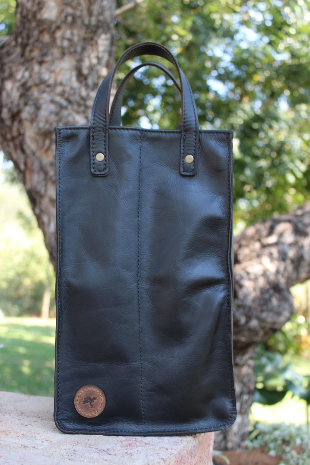 A black leather wine carrier with dual pockets and carry straps, displayed outdoors.