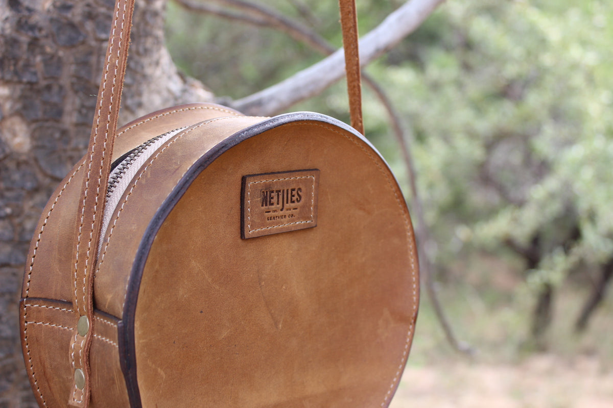 Round, brown leather shoulder bag hanging from a tree branch