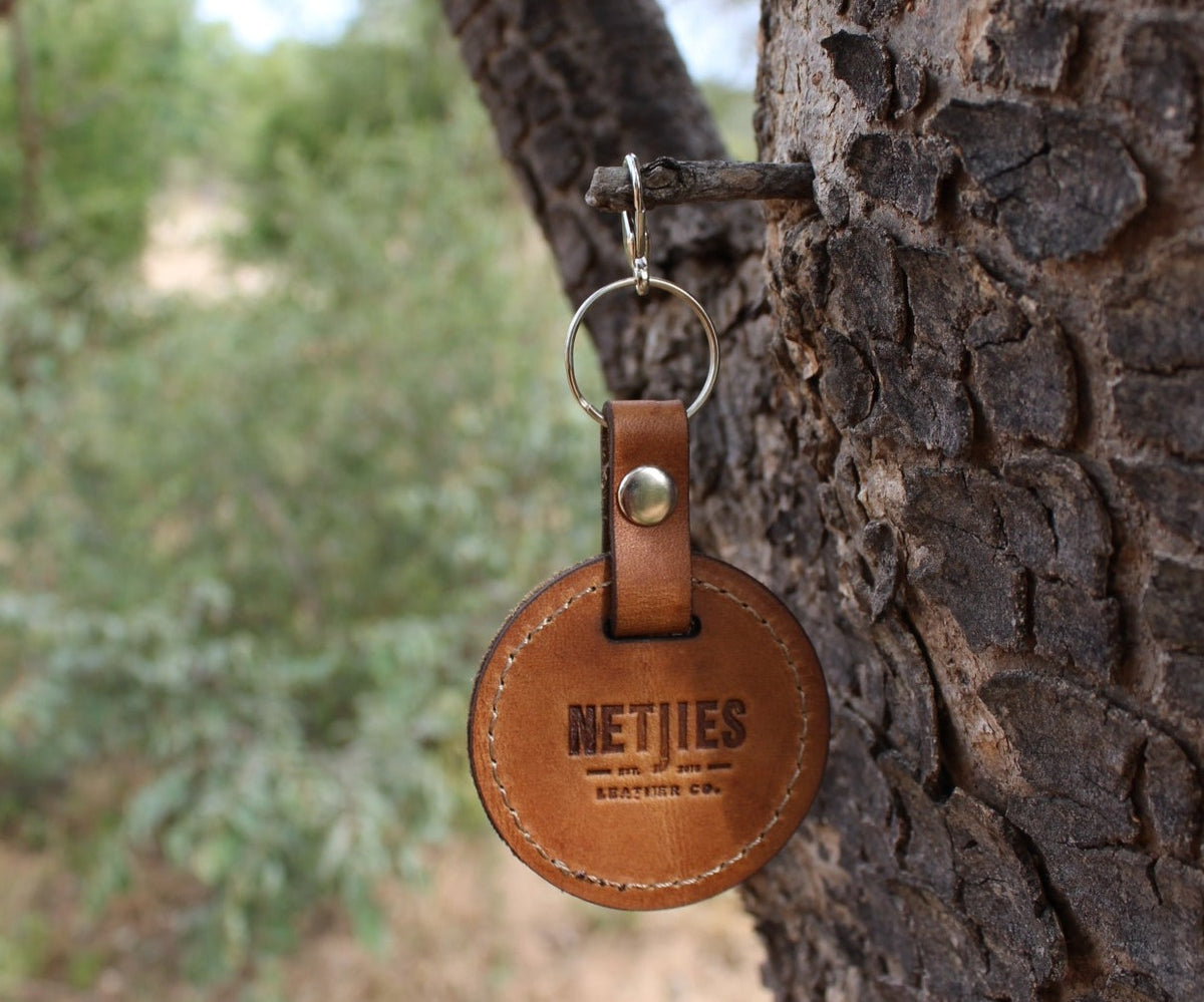 A brown leather keychain with a metal ring, featuring a warthog pattern, hanging from a tree branch.