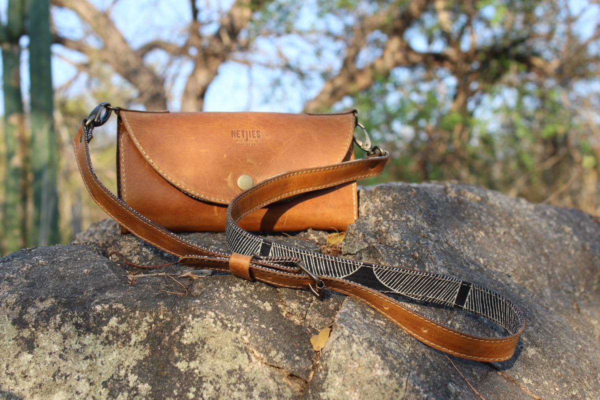 Handmade brown leather bag by Netjies Leather Co with a strap on a rock, blurred natural background.