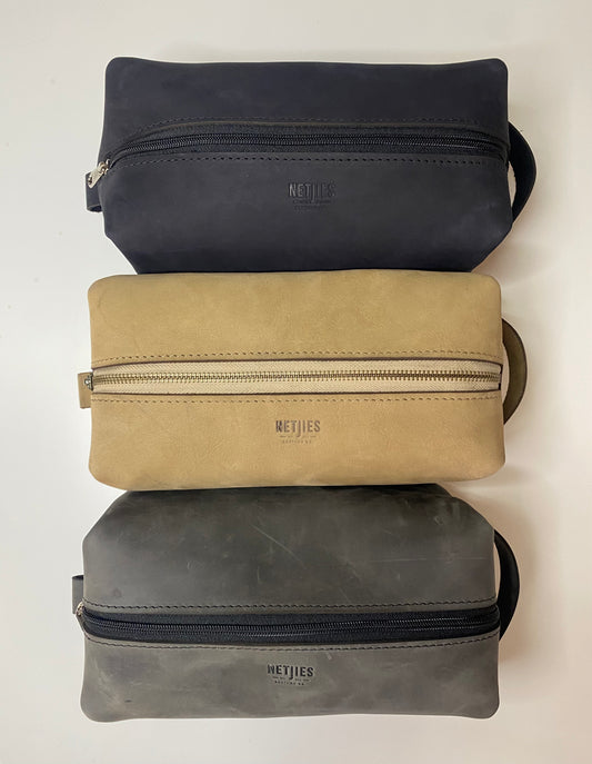 Three toiletry bags placed horizontal to each other on a surface. The top bag is black, the middle is beige, and the bottom one is gray.