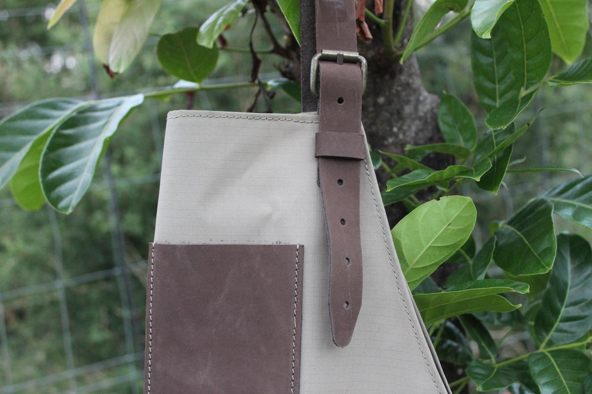 A beige canvas apron with brown leather details and a strap, displayed hanging on a tree surrounded by green leaves.