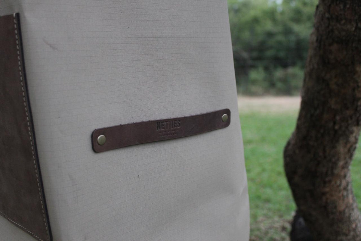 A beige canvas apron with brown leather details and a strap, displayed hanging on a tree surrounded by green leaves.