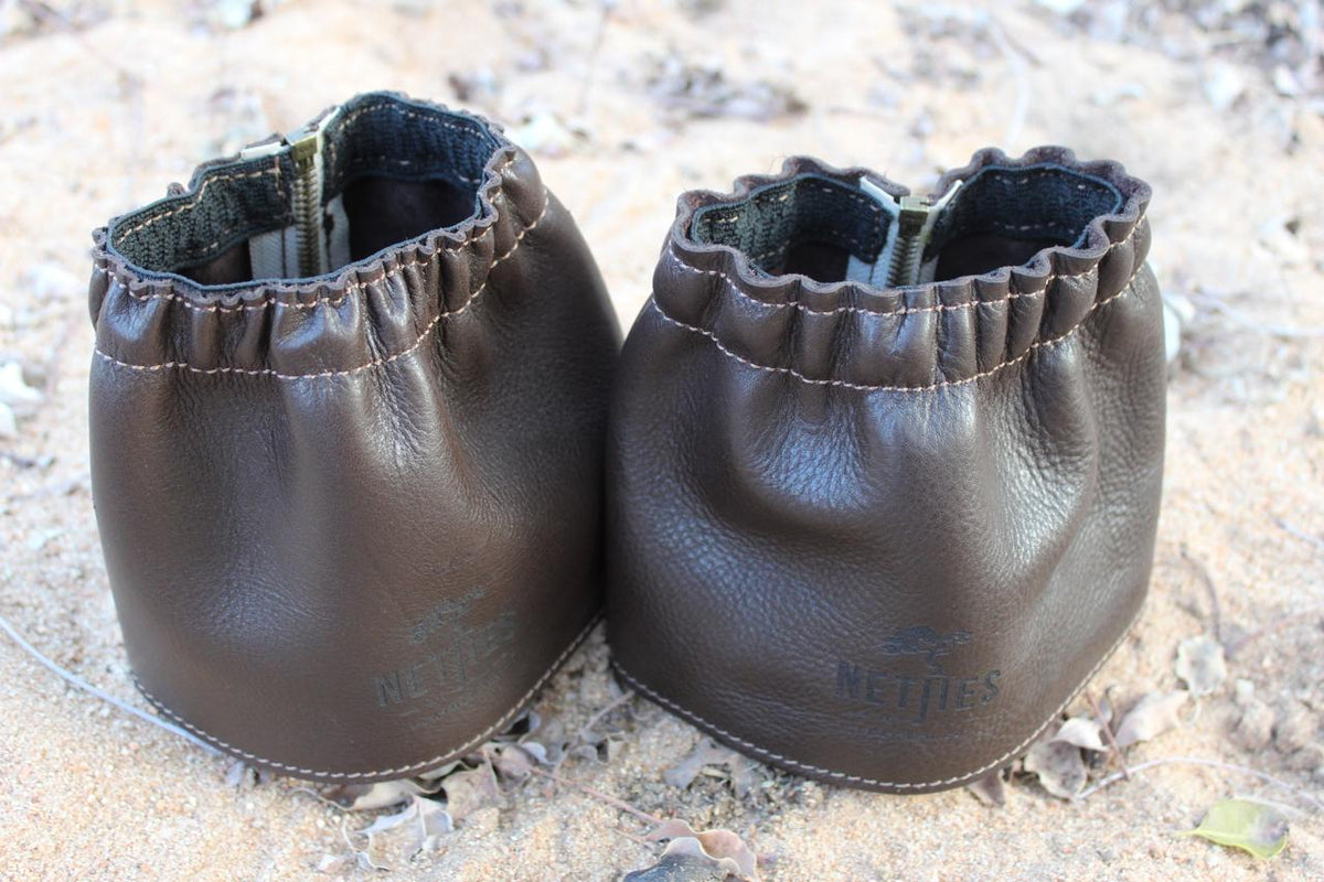 A pair of brown leather gaiters for shoes, displayed on a sandy surface.