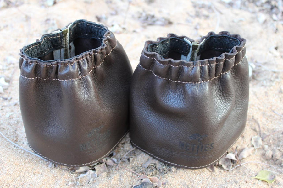 A pair of brown leather gaiters for shoes, displayed on a sandy surface.