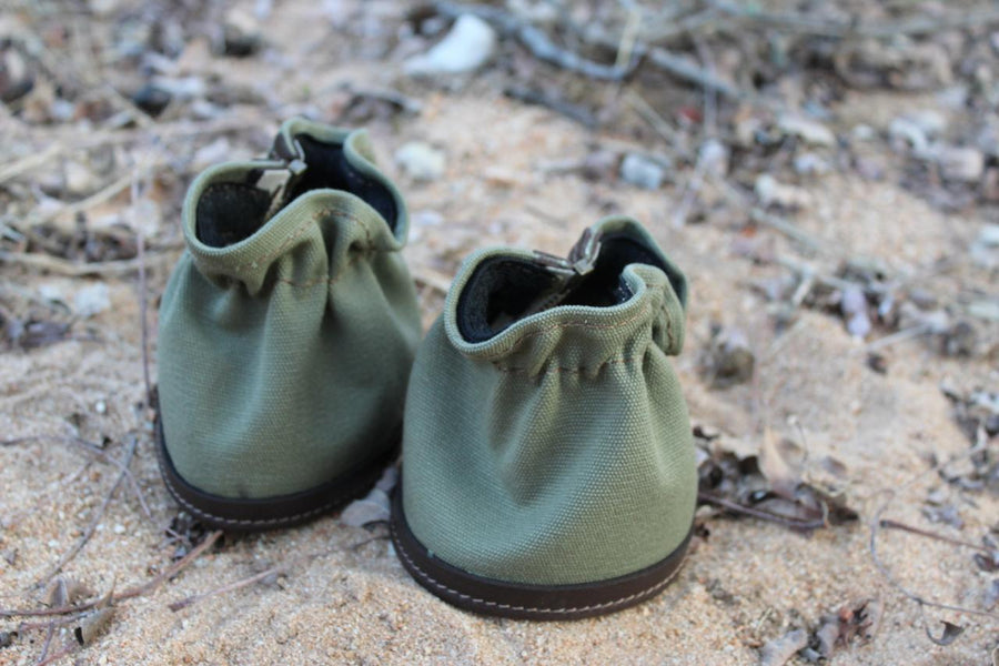A pair of green shoe gaiters placed on a sandy surface with natural surroundings.