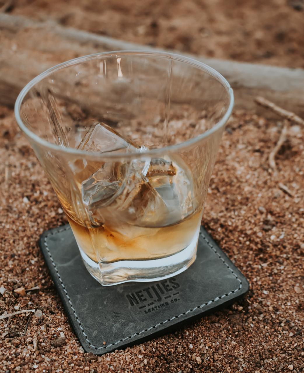 Glass of whiskey with ice on a leather coaster on a sandy surface