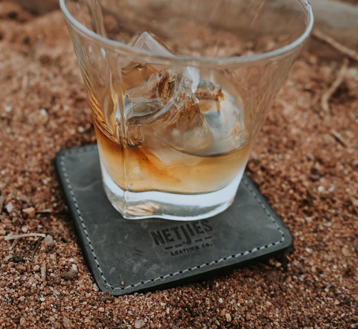 Glass of whiskey with ice on a leather coaster with 'NETJIES' branding, on a sandy surface.
