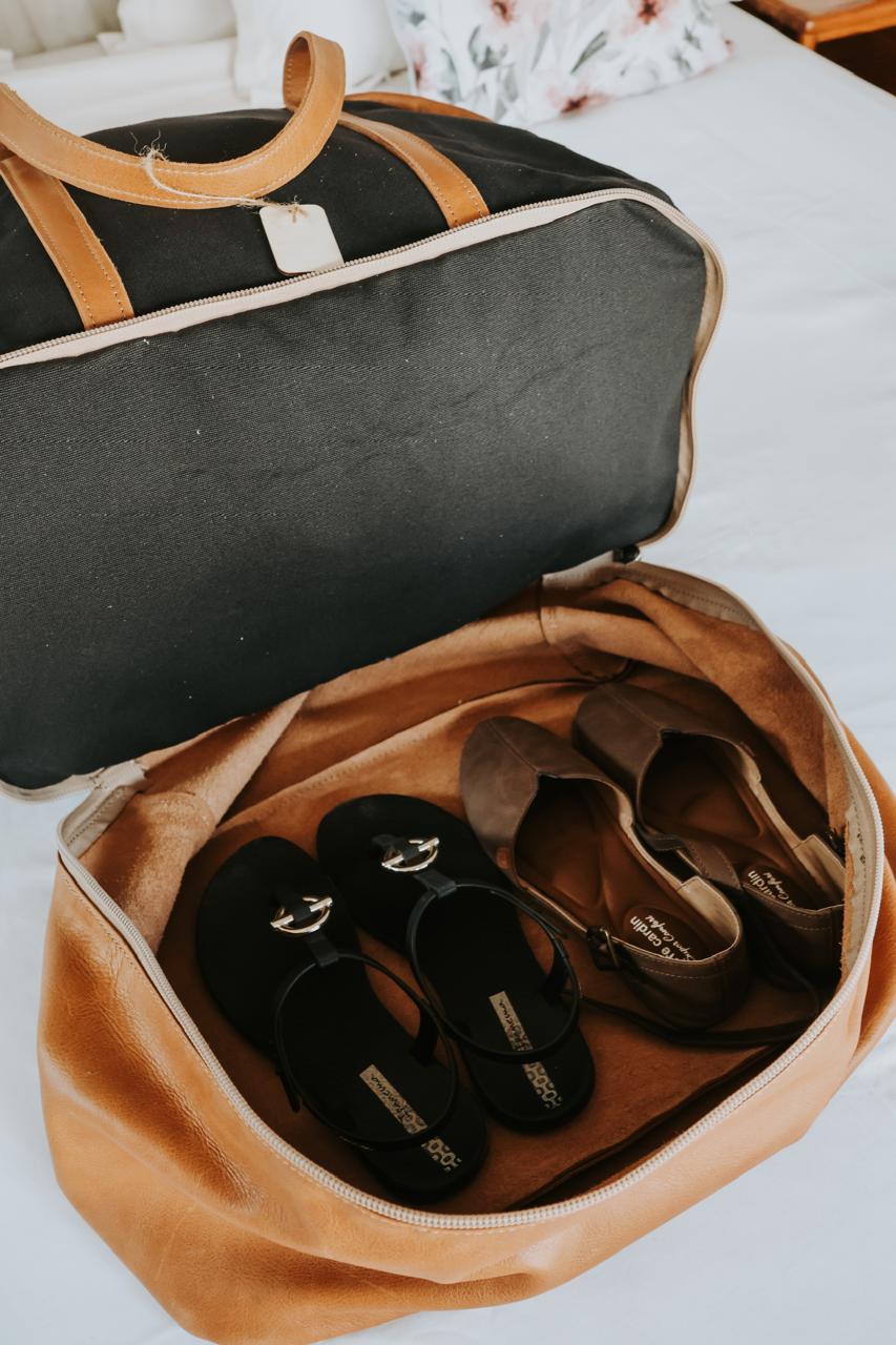 Open tan duffel bag with black sandals inside on a white surface