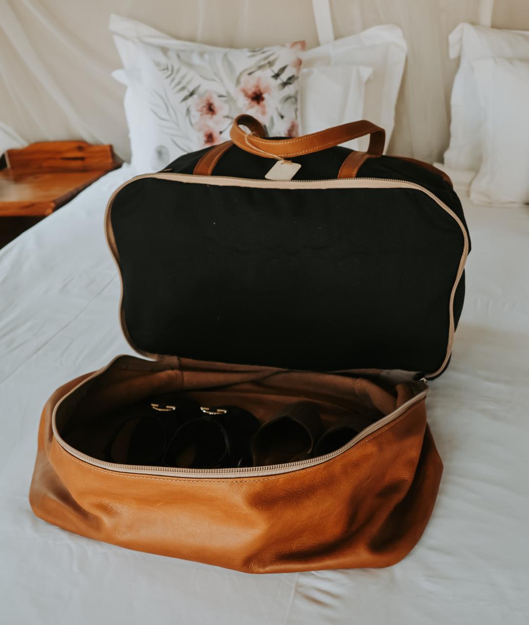 Brown and black duffel bag on a bed with pillows in the background