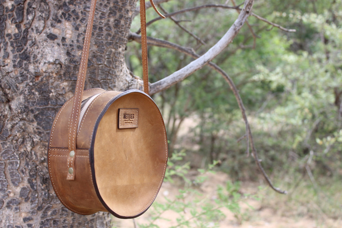 Round, brown leather shoulder bag hanging from a tree branch