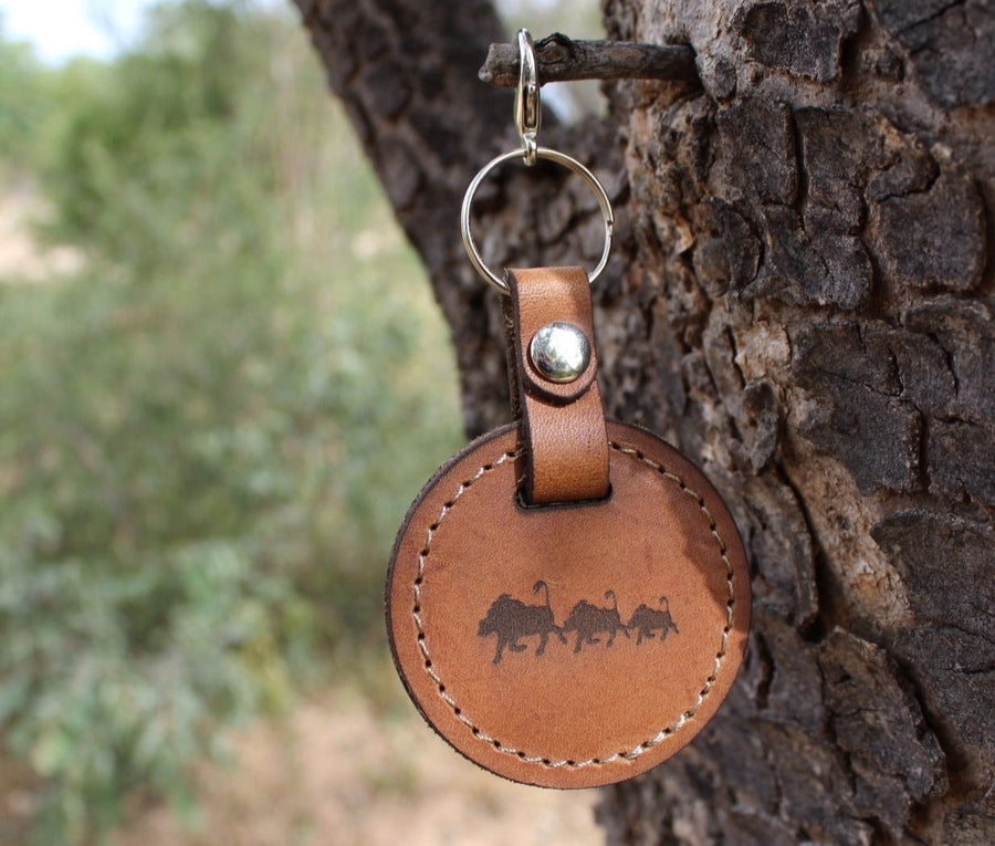 A brown leather keychain with a metal ring, featuring a warthog pattern, hanging from a tree branch.
