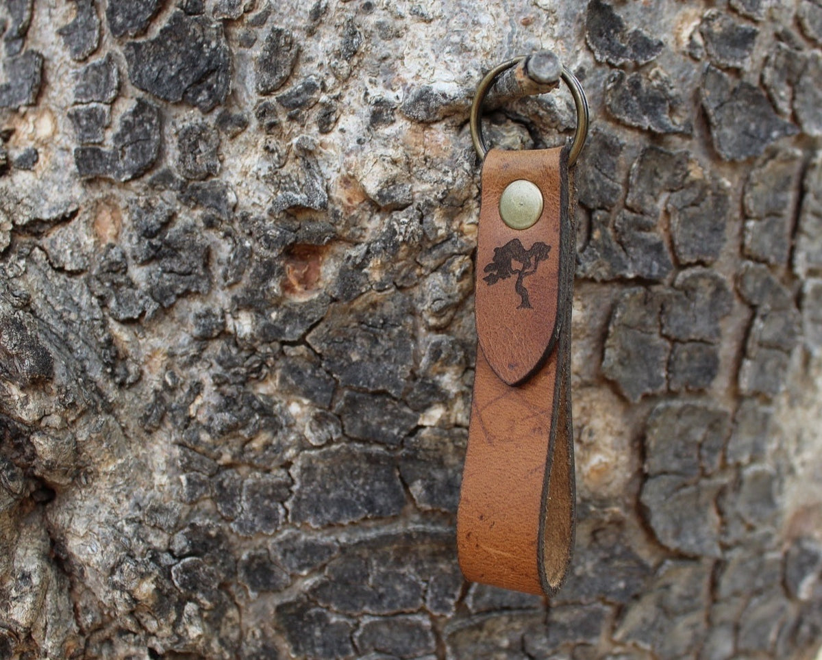 A brown leather keychain with a metal ring and a stamped tree logo, attached to a textured tree bark.
