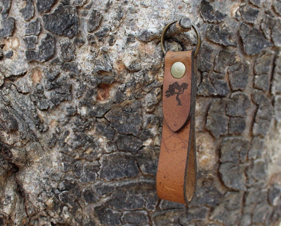 A brown leather keychain with a metal ring and a stamped tree logo, attached to a textured tree bark.