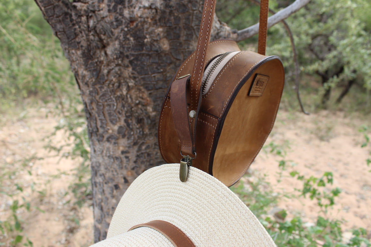 A brown leather hat clip attached to a straw hat, which is hanging on a tree.