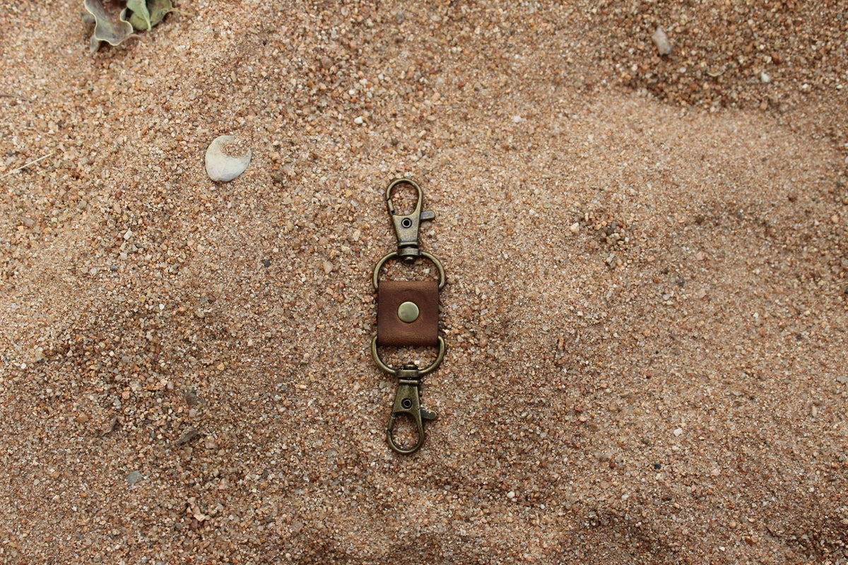 A metal key clip with a leather strap, placed on a sandy surface.