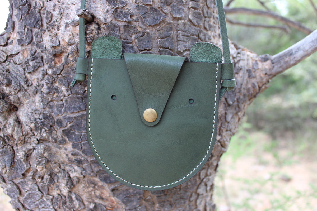 A green leather handbag with a flap closure and a metal snap button, displayed on a tree.